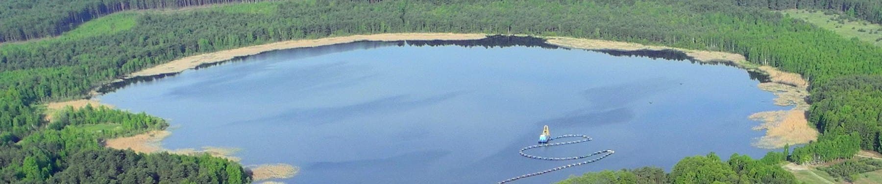 Aerial view of Lake Prybich in Ukraine, with a floating dredger and a curved line of pipeline floats on open water ringed by forest